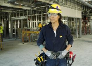 A woman in hard hat, toolbelt and protective goggles stands confidently in a building undergoing renovation.