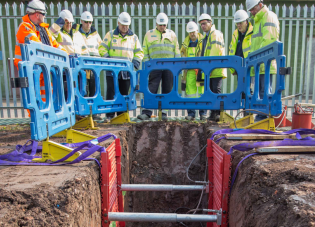 A group of workers in high visibility clothing and hard hats stand around a trench that has protective barriers preventing them from falling in as well as shoring inside to keep it from collapsing.