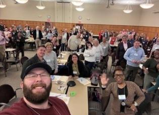 A group of people gathered around tables in a large room smile for a selfie-style photo.