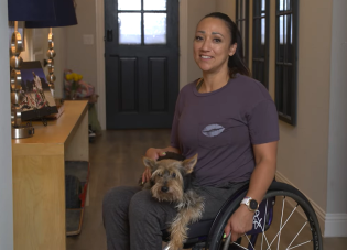 Vanessa Ross at home. She uses a wheelchair and is holding a small dog.