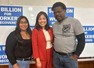 A woman with long dark hair wearing a black shirt and jeans stands with a woman with collar-length black hair wearing a red suit and a man with short dark hair wearing a grey t-shirt and jeans. They are smiling for a photo and standing in front of a white board with signs that read, " $1 BILLION FOR WORKERS RECOVERED."