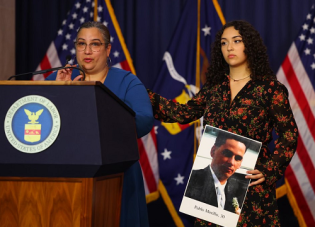 Wanda Engracia speaks at a podium. Her 18-year-old daughter Samatha stands beside her, holding a photo of her deceased father, with a hand on her mother's shoulder.