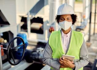 A middle-aged Black woman in a suit, safety vest, mask and hardhat poses at an industrial site.