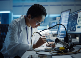 A woman sits at a desk in a lab coat working on a piece of computer equipment using a magnifying glass