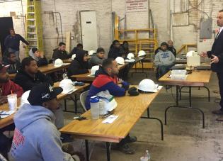 A man wearing a suit addresses a room full of apprentices. They sit at folding tables with their hard hats and other work gear.