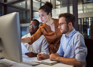 Three workers lean over a desk, looking at a computer screen.