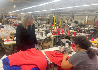 In a crowded workroom, a woman pushes red fabric through a sewing machine.