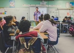 A teacher stands in a classroom addressing a dozen teenagers seated at desks with backpacks on the chairs.