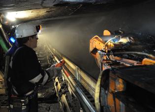 A coal miner wearing a helmet with a headlamp controls a continuous mining machine underground.