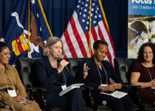 A seated woman in a business jacket addresses the audience. Three other women on the panel listen attentively.