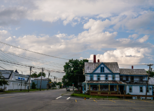 A quiet street scene in a small rural town showing an older two-story house with weathered blue and white paint under a partly cloudy sky, with electrical lines crisscrossing above.