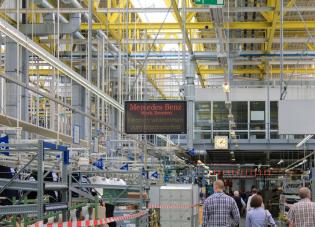 Several people in plain clothes walk through an auto factory. A sign in Germany reads “Mercedes-Benz Bremen plant, welcome to the employee party.”