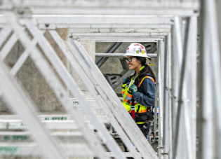 A woman in a hardhat and protective gear stands on scaffolding, overlooking a construction site.