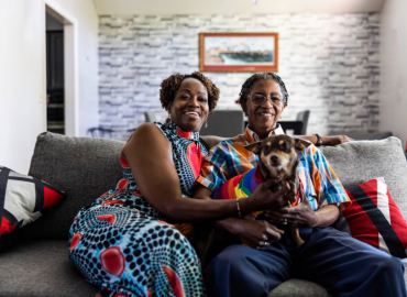 两个人面带微笑，坐在舒适客厅的沙发上，抱着一只围着彩虹围巾的小狗。房间里装饰着砖墙和现代家具。