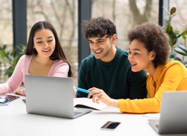 Tres jóvenes trabajan con una computadora portátil y cuadernos en una mesa, sonrientes y concentrados en su tarea. El escenario parece ser una habitación luminosa con grandes ventanales.