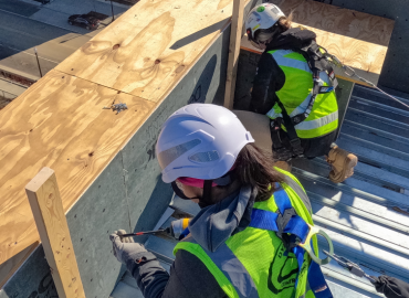 Dos empleadas de la construcción que usan equipo de protección contra caídas, incluidos cascos de seguridad, arneses y otros equipos.