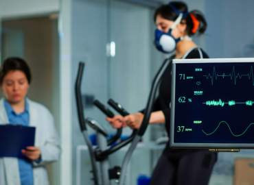 An exercise physiologist takes notes while collecting data from a woman on an elliptical machine who is wearing a respirator.