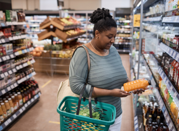 Una mujer con la cesta de compras en la mano mira una botella de jugo en la heladera de un pasillo del supermercado.