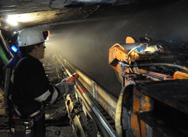 A coal miner wearing a helmet with a headlamp controls a continuous mining machine underground.
