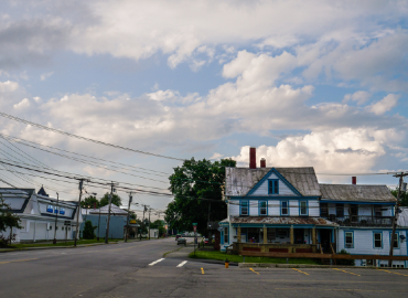 Una tranquila escena callejera en un pequeño pueblo rural que muestra una casa antigua de dos plantas con pintura azul y blanca desgastada bajo un cielo parcialmente nublado, con cables de electricidad que se entrecruzan por encima.