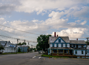 A quiet street scene in a small rural town showing an older two-story house with weathered blue and white paint under a partly cloudy sky, with electrical lines crisscrossing above.