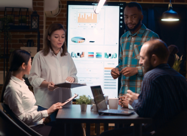 Varios empleados trabajan alrededor de una mesa con computadoras portátiles y tabletas. Detrás de ellos, hay una gran pantalla con una presentación sobre datos.